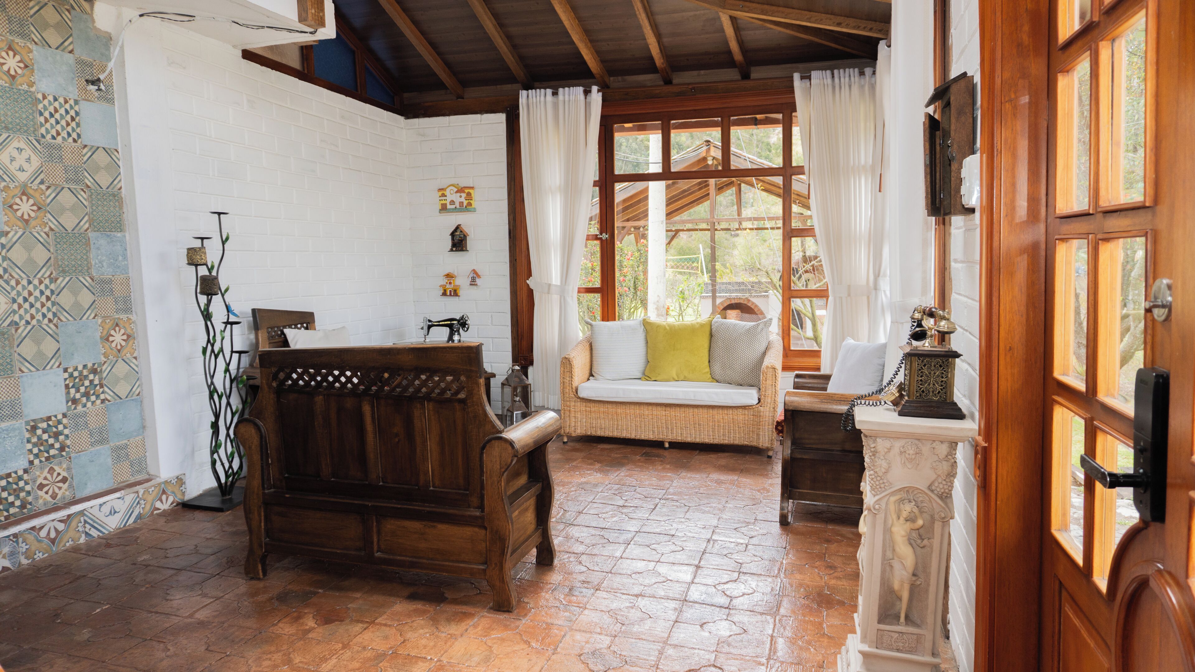 A room with a wooden bench, a wicker sofa, a white brick wall, and a window with curtains.