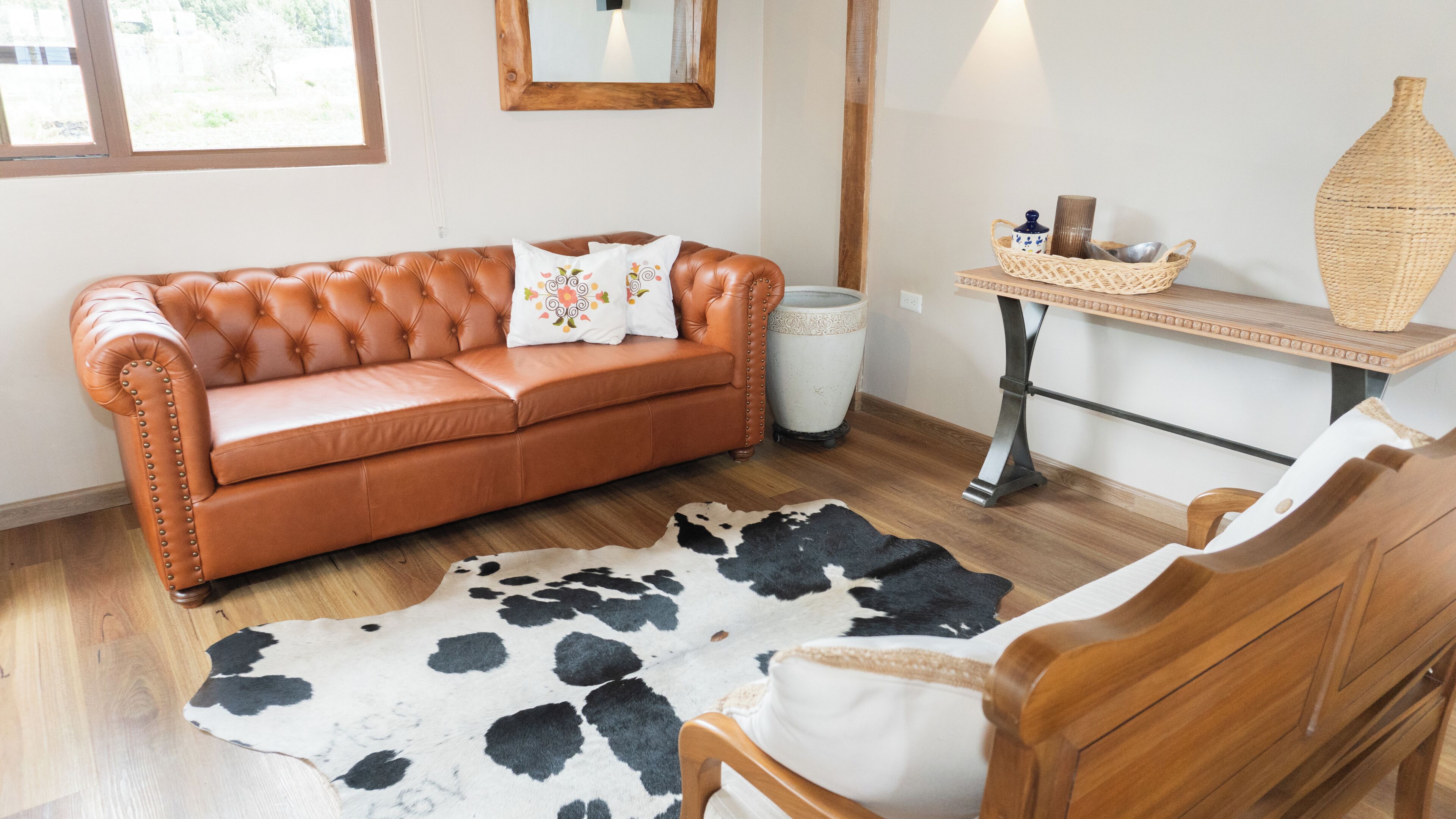 A room with a leather sofa, a cowhide rug, a wooden chair, a small table with a woven basket, and a vase.