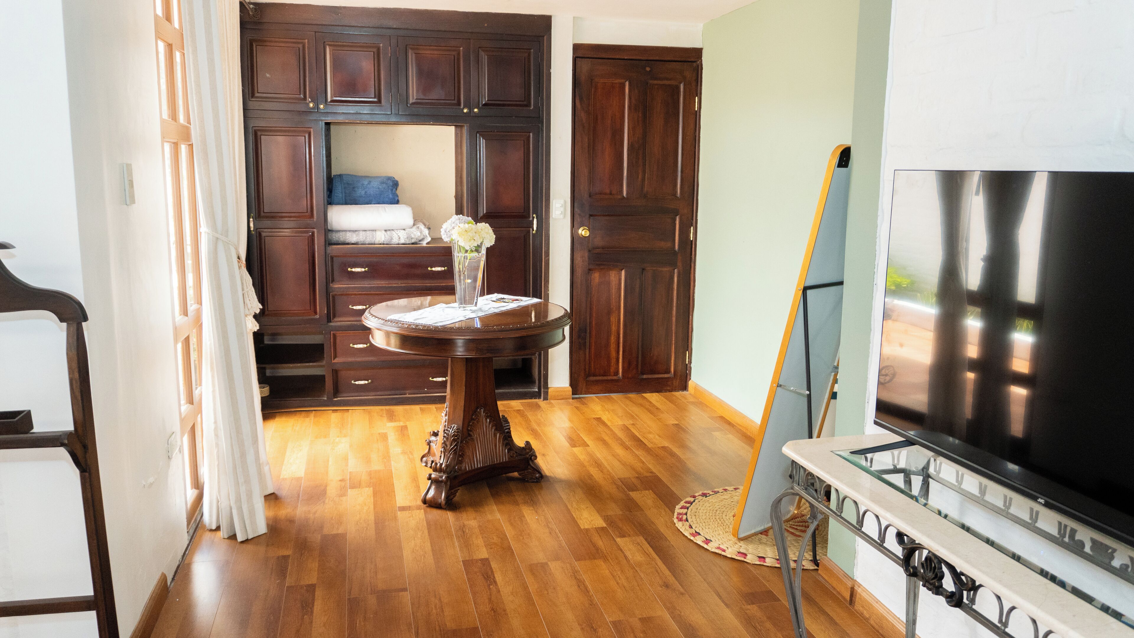 A room with a wooden floor, a round table with a vase, a large dark TV, and a wardrobe.