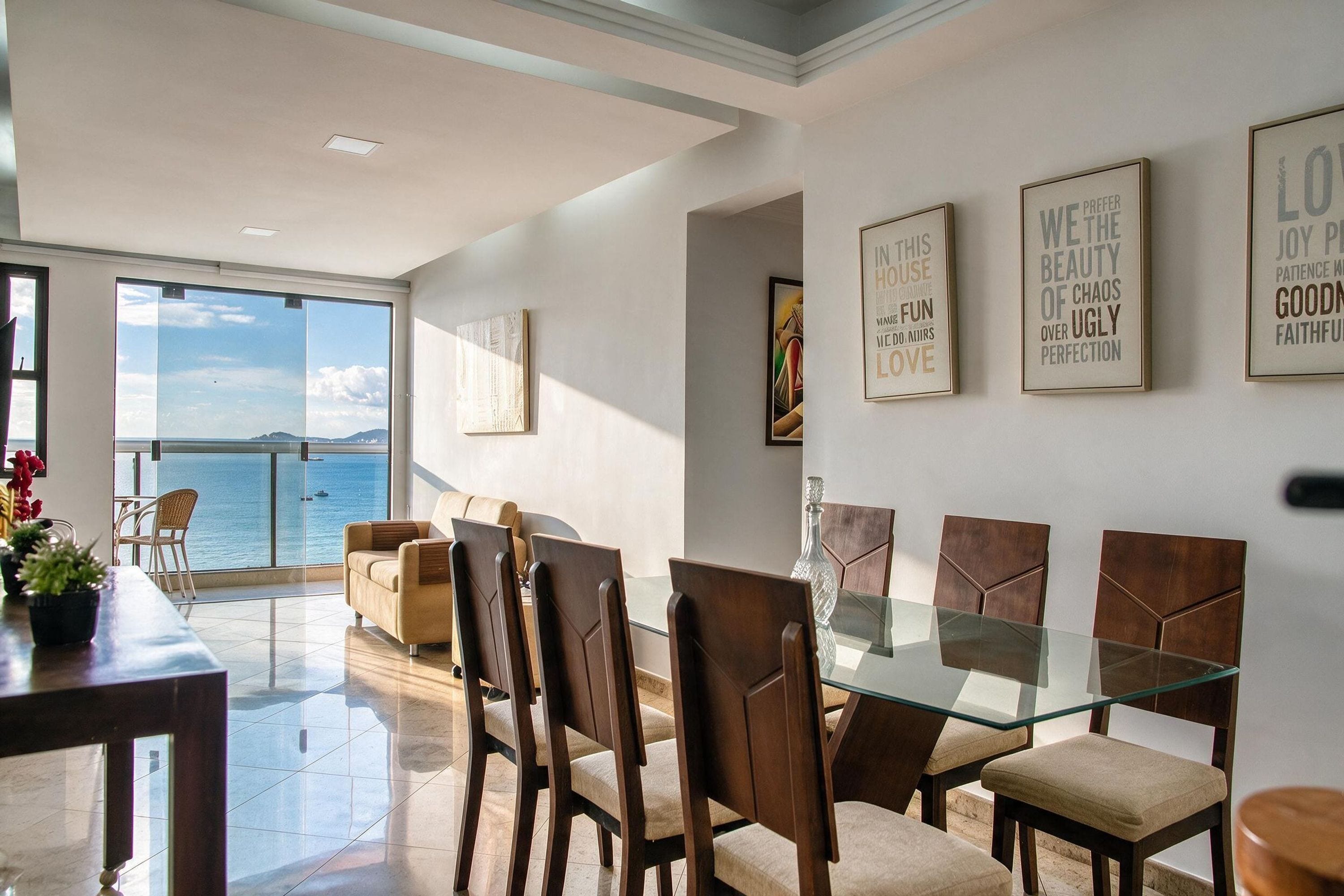 Un espace repas avec une table et des chaises en verre, une vue sur la mer grâce à de grandes fenêtres et des œuvres d’art murales inspirantes.
