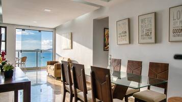 Uma sala de jantar com mesa e cadeiras de vidro, vista para o mar através de grandes janelas e quadros com mensagens inspiradoras.