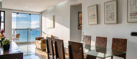 A dining area with a glass table and chairs, a view of the sea through large windows, and motivational wall art.