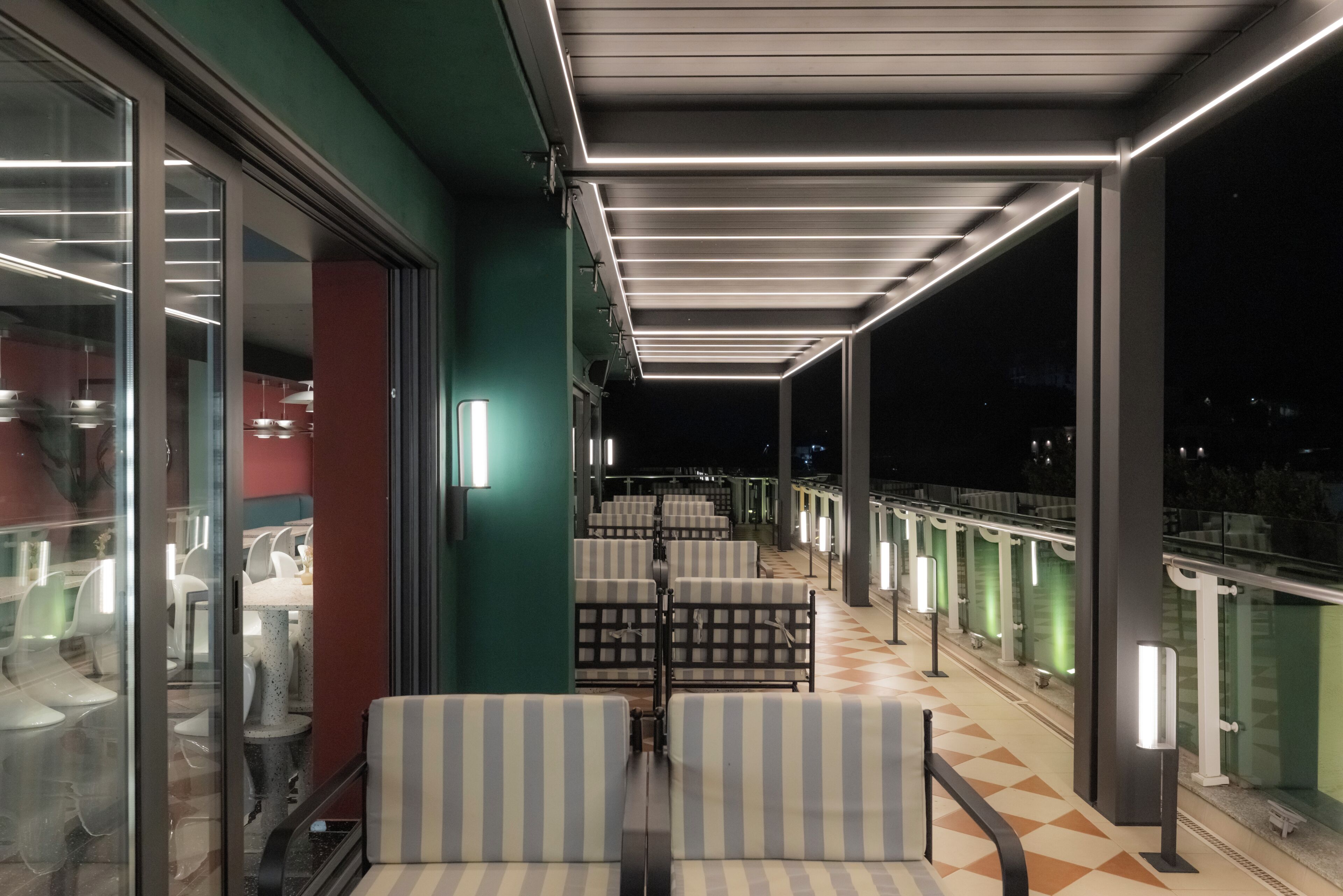 A covered outdoor seating area with striped chairs and tables, a glass railing, and a view of the night sky.