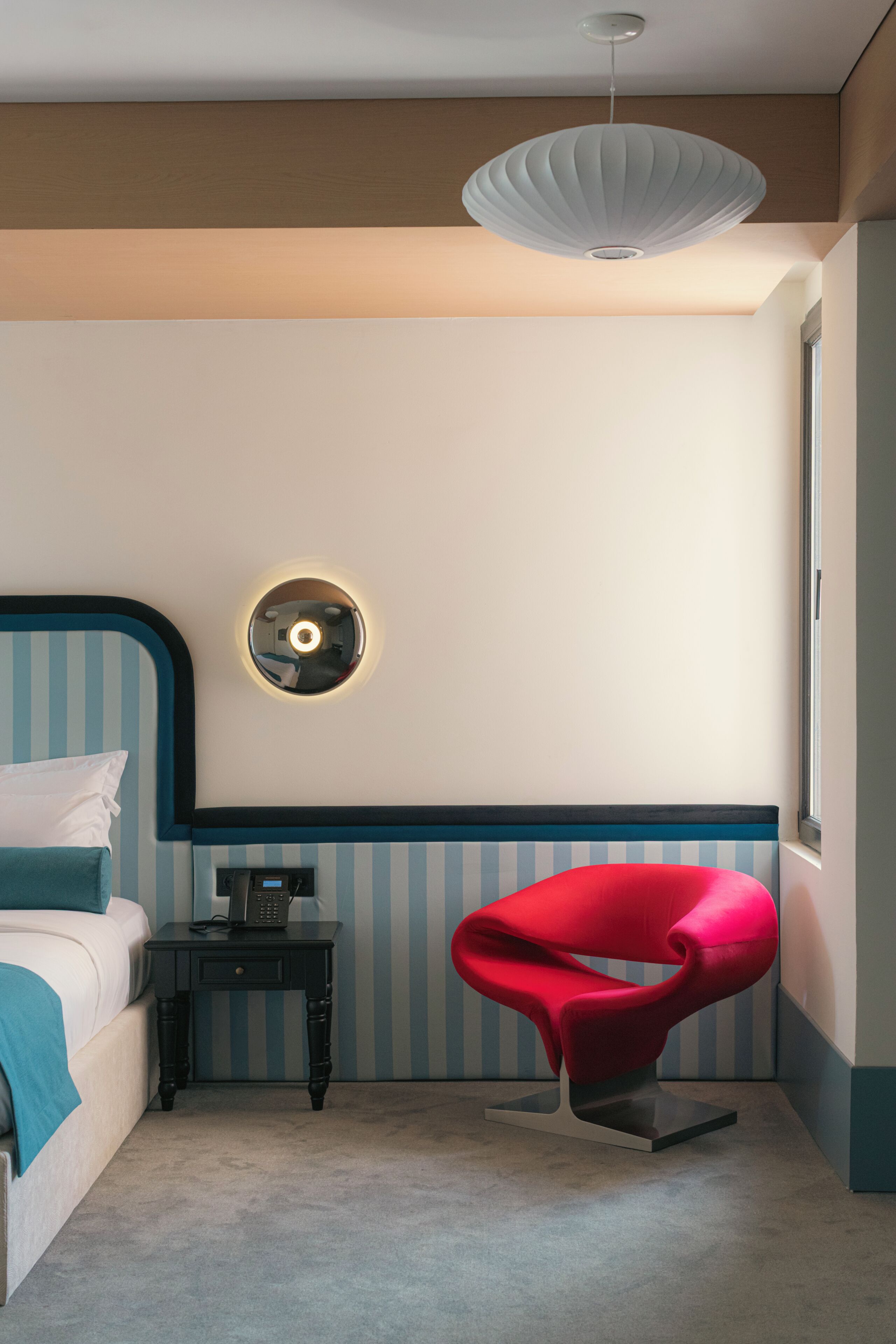 Une chambre à coucher avec un lit, une table de chevet, une chaise rouge, une lampe fixée au mur et une fenêtre.