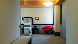 A hotel room with a bed, a bench, a red chair, and a small table.