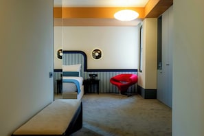 A hotel room with a bed, a bench, a red chair, and a small table.