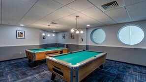 Sala com mesa de bilhar, duas mesas de bilhar, luminárias no teto, quadros nas paredes e um espelho circular.