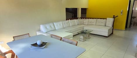 Un comedor con mesa blanca y sillas de madera, sofá blanco y una pared de acento amarilla.
