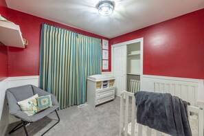 Ein Zimmer mit roter Wand, einem weißen Heizkörper, einem grauen Sessel mit Kissen, einem gestreiften Vorhang und einer weißen Tür, hinter der sich ein weiterer weißer Heizkörper befindet.