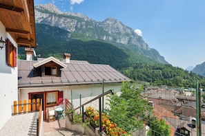 A mountain cabin with a sloped roof, a small porch with a table and chairs, and a view of a valley with buildings and mountains in the background.