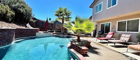 Una piscina con borde de ladrillo, rodeada de un patio con sillones de descanso y una palmera.