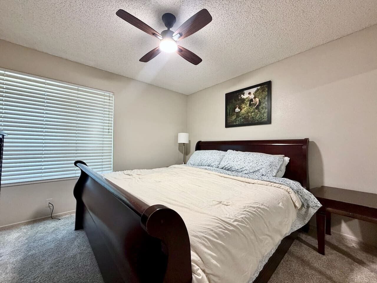 Un dormitorio con cama, ventilador de techo, ventana con persianas, mesita de noche y lámpara de pared.