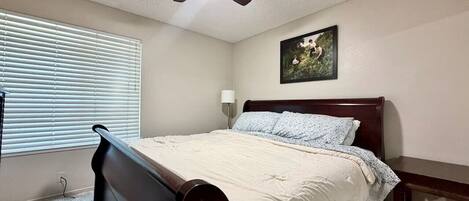 Un dormitorio con cama, ventilador de techo, ventana con persianas, mesita de noche y lámpara de pared.