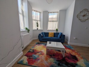 Ein Wohnzimmer mit einem blauen Sofa, einem weißen Couchtisch und einem farbenfrohen Teppich. Zwei Fenster mit Spitzenvorhängen und Blumentöpfen sind zu sehen.