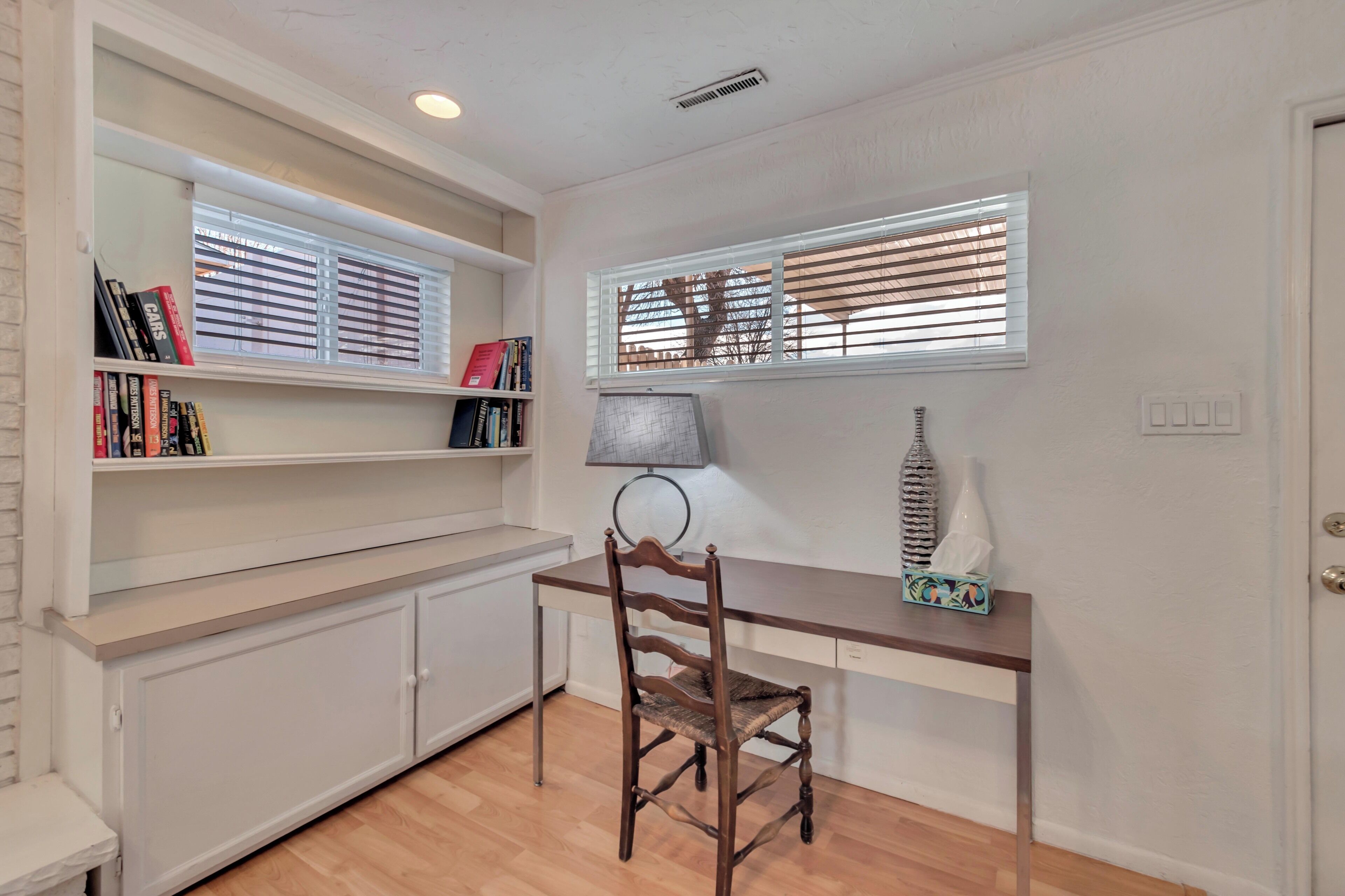 Una habitación con escritorio de madera, una silla, una estantería y un ventanal con persianas.