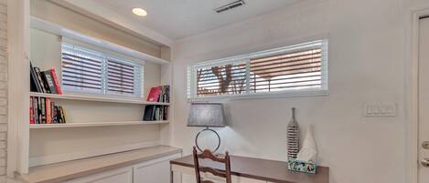 Una habitación con escritorio de madera, una silla, una estantería y un ventanal con persianas.
