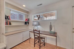 Ein Zimmer mit Schreibtisch aus Holz, einem Stuhl, einem Bücherregal und einem Fenster mit Jalousien.
