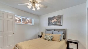 Un dormitorio con cama, mesita de noche, ventilador de techo, ventana con persianas y un cuadro en la pared.
