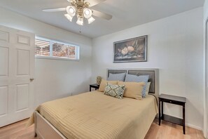 Ein Schlafzimmer mit Bett, Nachttisch, Deckenventilator, Fenster mit Jalousien und einem Gemälde an der Wand.