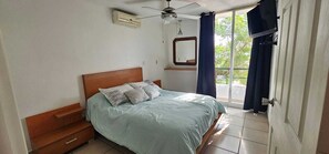 Un dormitori amb un llit, dues taules de nit de fusta, un ventilador de sostre, una finestra amb cortines i una porta.