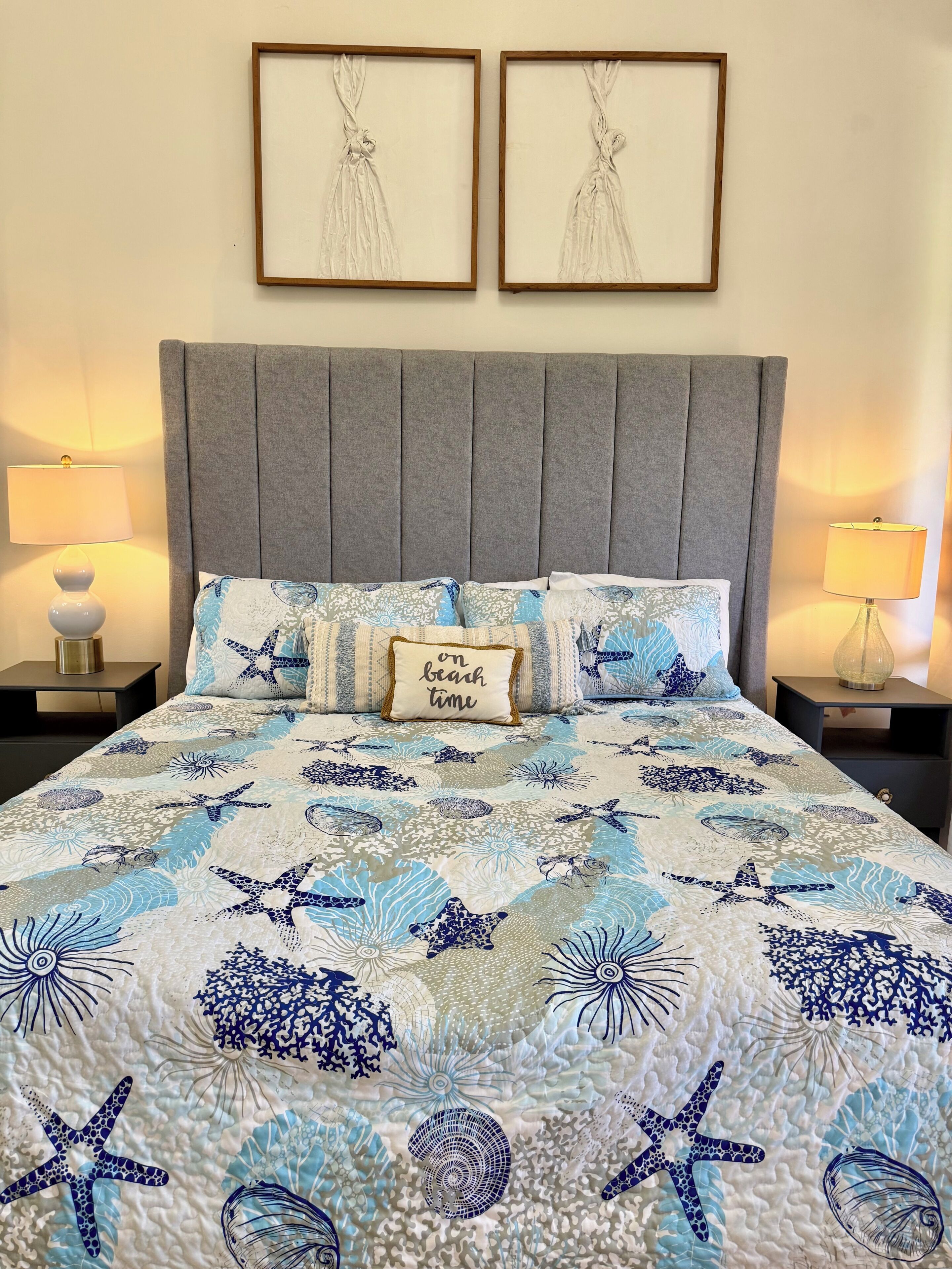 Una cama con colcha temática oceánica en tonos azul y blanco, dos cuadros enmarcados en la pared y dos lámparas de noche.
