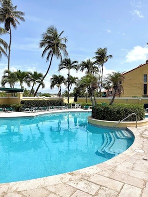 A swimming pool with lounge chairs, palm trees, and a building with a red roof.