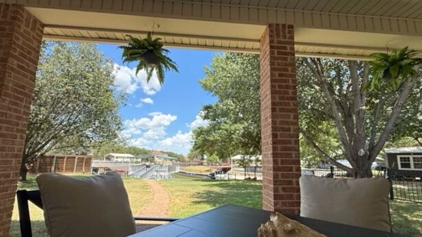 An outdoor patio with a table and chairs, a brick wall, and a view of a grassy area with trees and a distant building. - Oasis at Eagle Mountain Lake (Azle)
