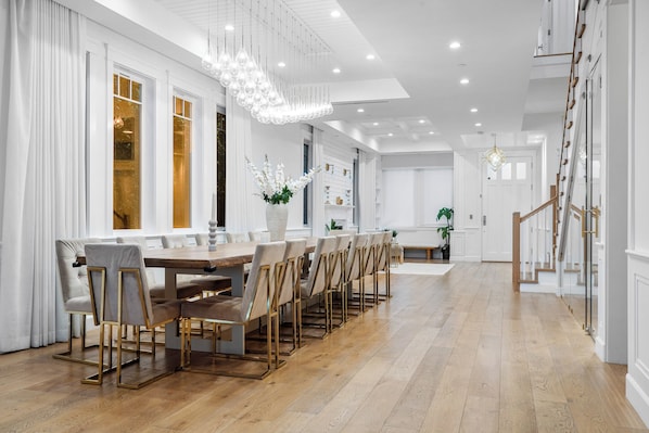 A spacious dining area with a long wooden table and multiple chairs, a staircase with glass railings, and a modern chandelier.
