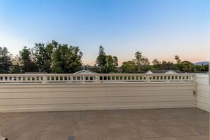 A flat roof terrace with a white railing, a tiled floor, and a backdrop of trees and buildings.
