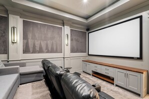 A home theater room with a large flat-screen TV, a leather recliner, grey sofas, and a wooden entertainment unit.
