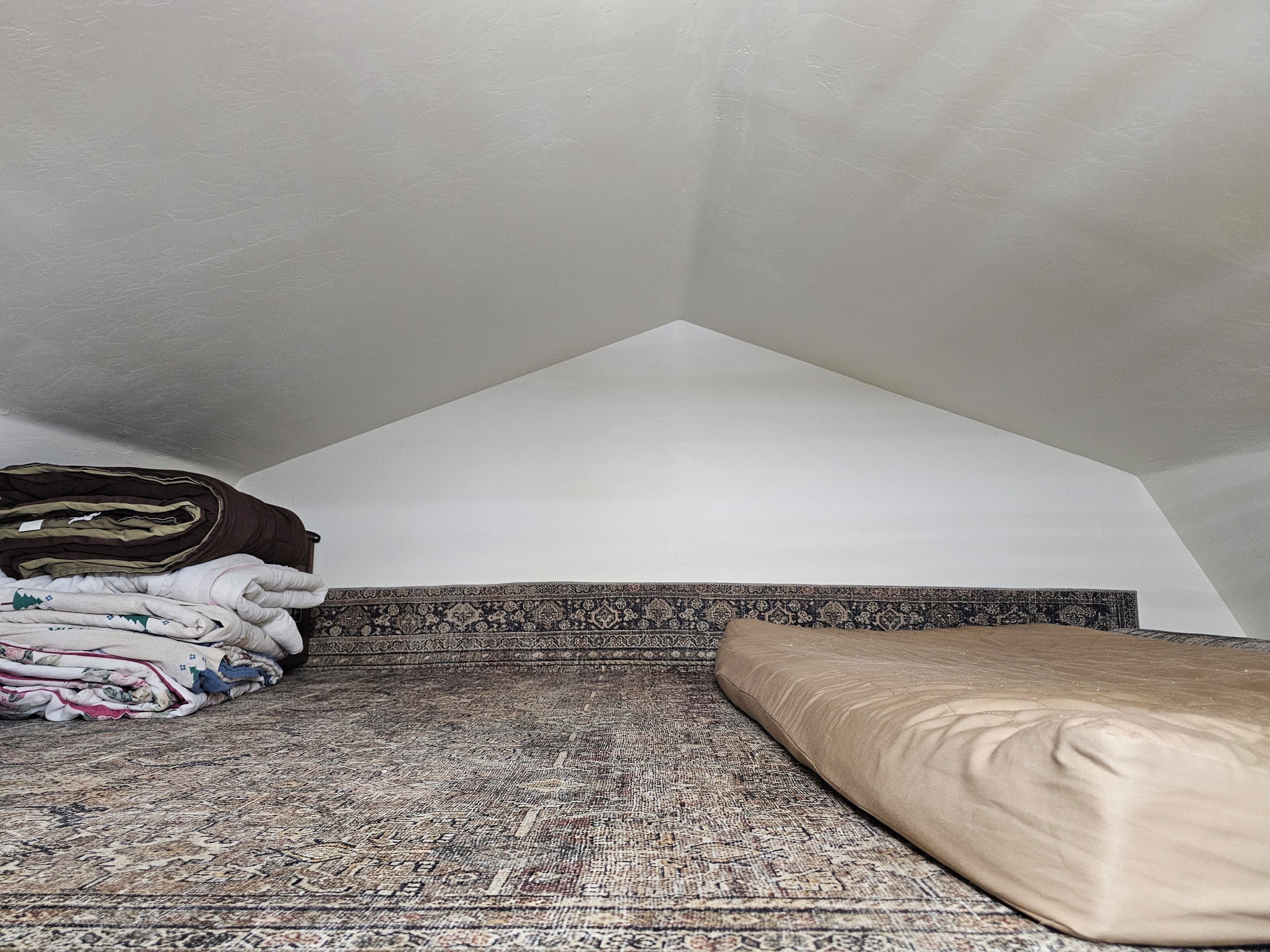 Une pièce avec un plafond en pente, un tapis à motifs, un lit avec un oreiller beige et des couvertures pliées sur le sol.