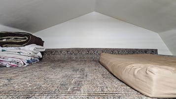 Une pièce avec un plafond en pente, un tapis à motifs, un lit avec un oreiller beige et des couvertures pliées sur le sol.