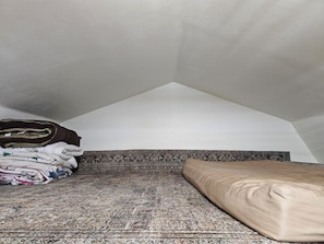 A room with a sloped ceiling, a patterned rug, a bed with a beige pillow, and folded blankets on the floor.