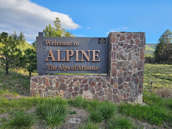A welcome sign for ALPINE - The Alps of Arizona, featuring a stone wall and a clear sky.