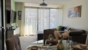 Zona pranzo con tavolo rotondo in vetro apparecchiato per quattro persone, un divano sullo sfondo e vista sulla città attraverso tende leggere.