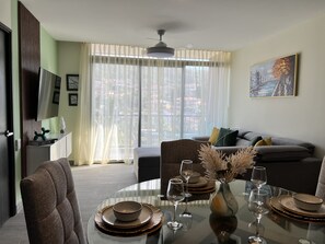 A dining area with a round glass table set for four, a sofa in the background, and a cityscape view through sheer curtains. - Modern 1-bedroom apartment in wonderful San Salvador with Pool & Gym (San Salvador)