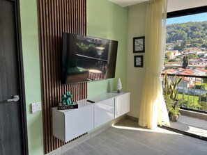 A modern living room with a flat-screen TV mounted on the wall, a white cabinet below, and a balcony with a view of a residential area. - Modern 1-bedroom apartment in wonderful San Salvador with Pool & Gym (San Salvador)