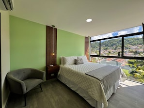 A bedroom with a bed, a chair, a nightstand, a window with a view of a town, and a wall with vertical lines. - Modern 1-bedroom apartment in wonderful San Salvador with Pool & Gym (San Salvador)