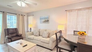 Ein Wohnzimmer mit einer Couch, einem Couchtisch, einem Esstisch mit Stühlen, einem Deckenventilator und einem Fenster mit Vorhängen.