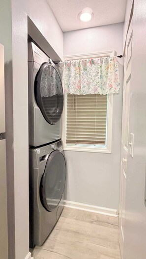 A laundry room with two stacked washing machines, a window with blinds, and a door with a light switch. - Modern Myrtle Beach Getaway|2BR Condo |Pool&Tennis (Myrtle Beach)