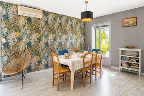 A dining area with a table set for four, surrounded by chairs, a hanging light fixture, and a bookshelf with books. - L'Ardèchoise - Piscine - Petfriendly (Vallon-Pont-d'Arc)