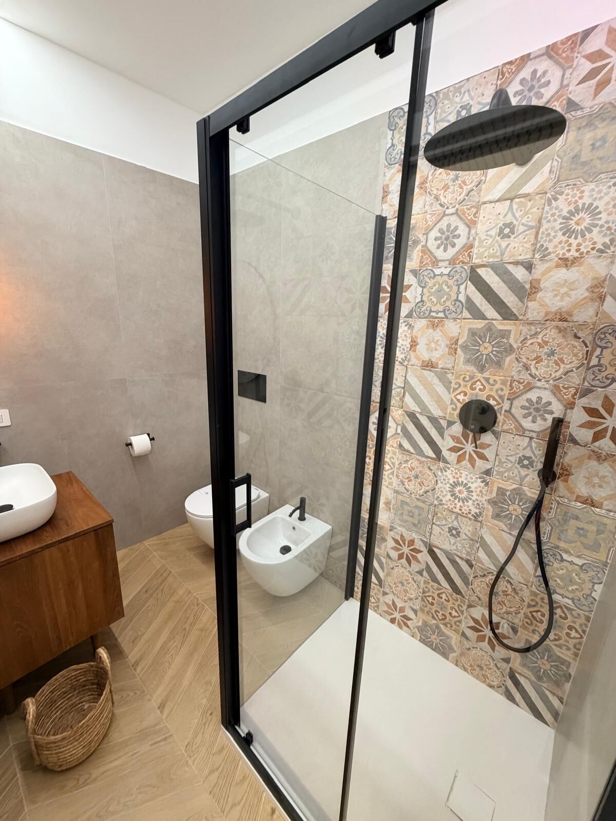 A bathroom with a glass shower enclosure, a white sink, a bidet, and a patterned tile wall.