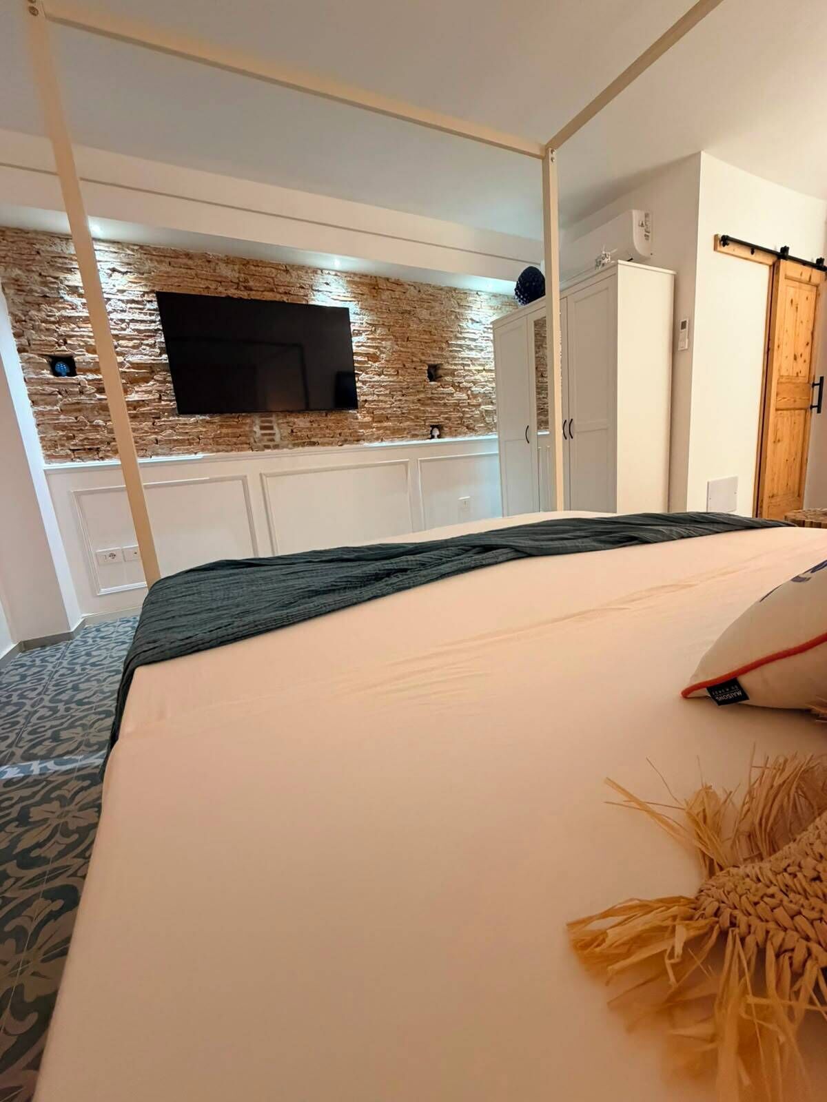 Une chambre d’hôtel comprenant un lit, un téléviseur fixé au mur, une armoire et un coin cuisine avec un mur en pierre.