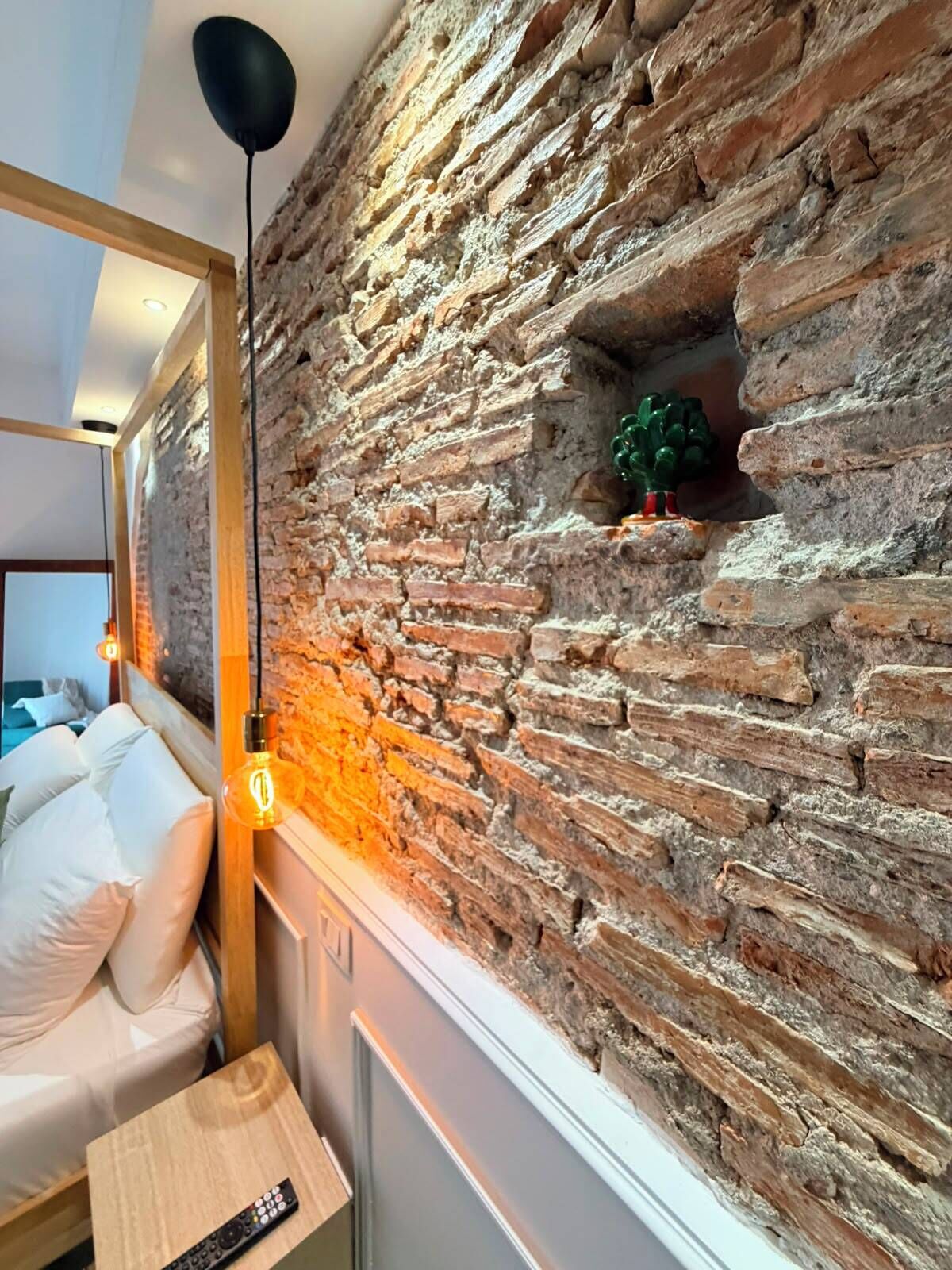 A room with a brick wall, a bed with white bedding, a wooden nightstand, and a hanging light fixture.