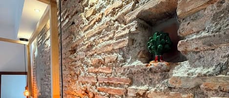 Una habitación con pared de ladrillo, cama con ropa de cama blanca, mesita de noche de madera y una lámpara colgante.