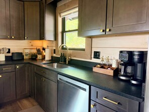 A modern kitchen with dark cabinetry, a stainless steel dishwasher, a coffee maker, and a window providing natural light. - New Cabin - Indoor Swim Spa, Hot Tub, Game Room! (Gatlinburg)