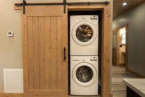 A laundry room with two stacked front-loading washing machines, wooden sliding doors, and a vent on the wall. - New Cabin - Indoor Swim Spa, Hot Tub, Game Room! (Gatlinburg)