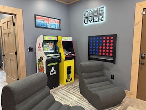 Ein Raum mit zwei Spielautomaten, einem an der Wand befestigten „Game Over“-Schild und zwei grauen Sesseln.