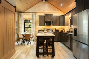 A modern kitchen with wooden cabinetry, stainless steel appliances, and a dining area with a round table and chairs. - New Cabin - Indoor Swim Spa, Hot Tub, Game Room! (Gatlinburg)