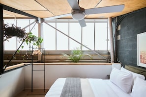 A bedroom with a bed, a ceiling fan, a window with plants, and a view of the city. - Bottega Loft Industrial Charm with Artistic Flair (Brunswick)